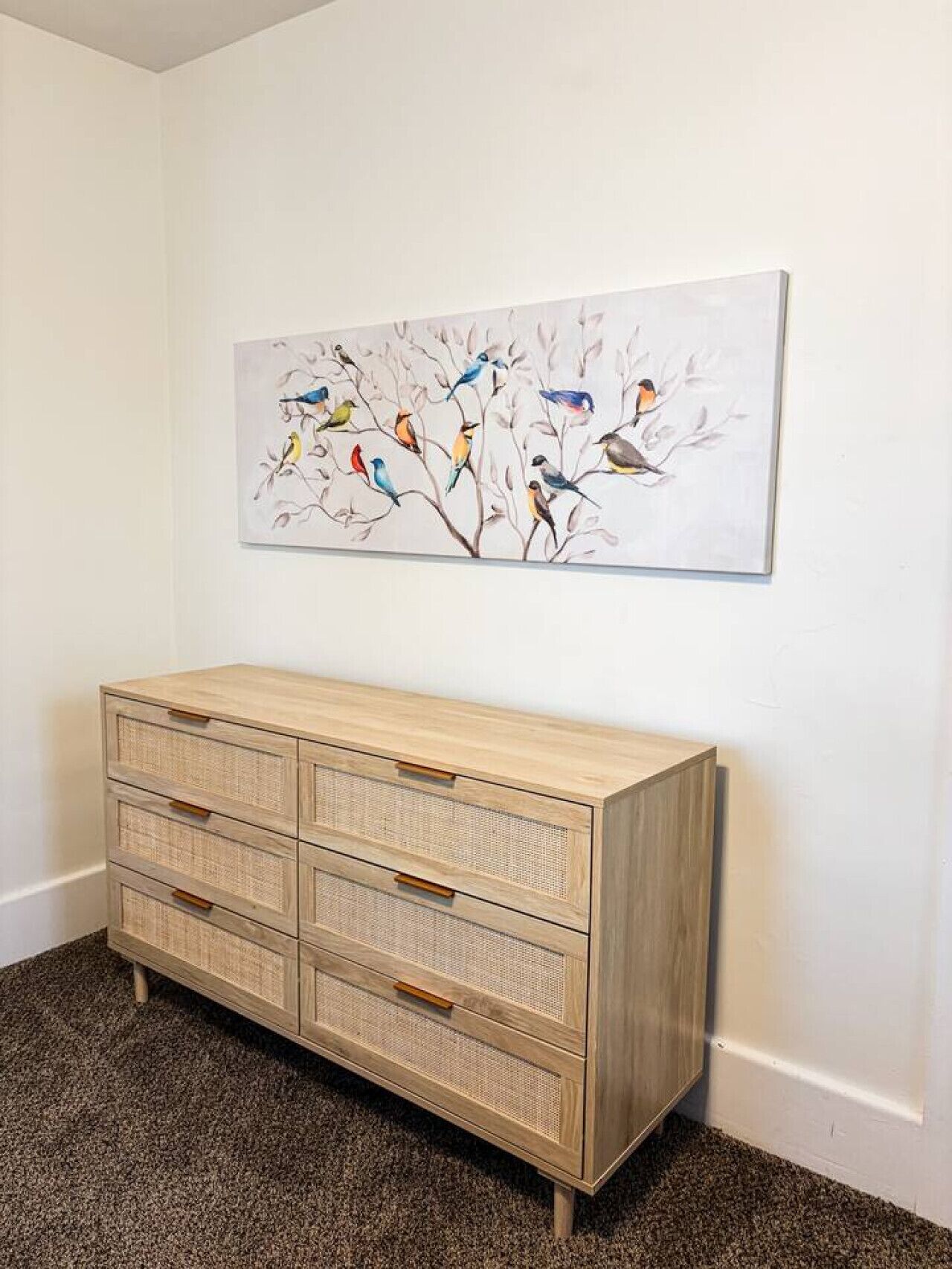 A room with a light wooden dresser, a wall-mounted painting of birds on branches, and a carpeted floor.