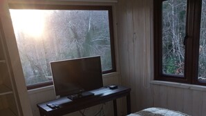 Ein Schlafzimmer mit einem Bett, einem Schreibtisch mit Computer und zwei Fenstern mit Blick auf Bäume.