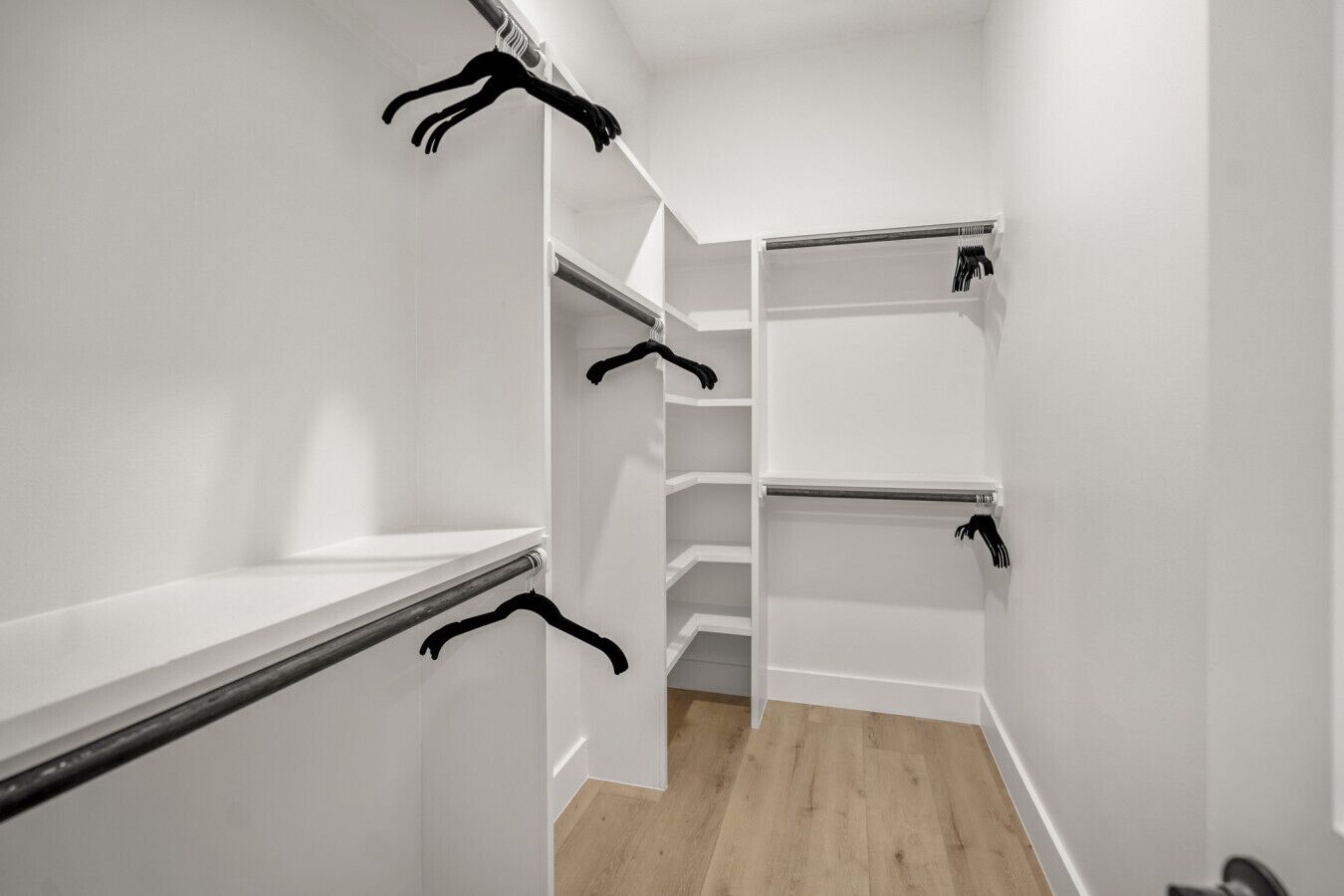 A walk-in closet with white walls, wooden flooring, and multiple shelves with black hangers.