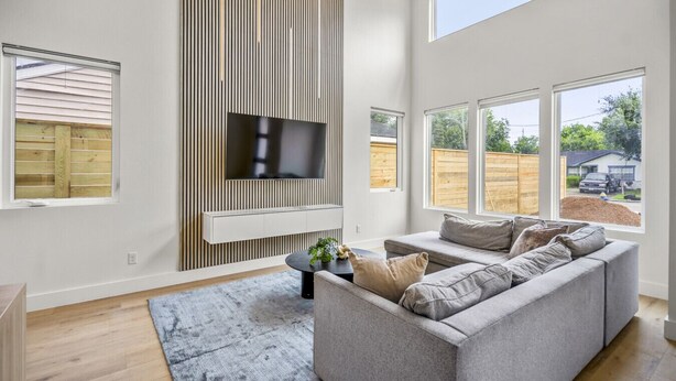 Ein modernes Wohnzimmer mit einem grauen Sofa, einem Flachbildfernseher an einer Wand aus Holzpaneelen und großen Fenstern mit Blick auf den Garten.