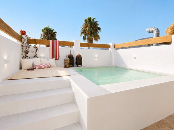 A rooftop terrace with a pool, white walls, wooden beams, and a seating area with cushions and a book.