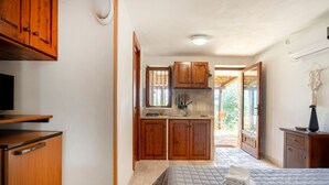 A bedroom with a bed, a small desk, and a wardrobe. There is a kitchen area with wooden cabinets and a window.