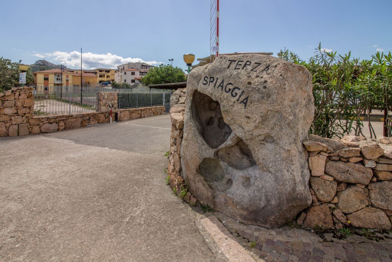 A stone marker with 'TERZA SPIAGGIA' inscribed, a paved road, and residential buildings in the background.