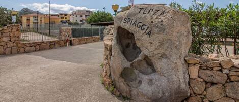 A stone marker with 'TERZA SPIAGGIA' inscribed, a paved road, and residential buildings in the background.