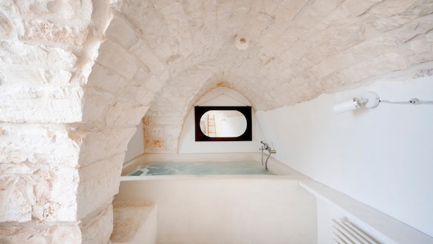 Un baño con paredes de piedra, una bañera, un ventanal pequeño y una l ámpara fijada a la pared.