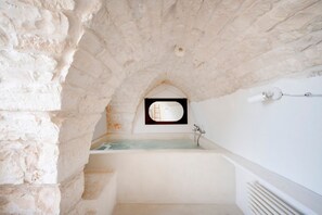 A stone-walled bathroom with a bathtub, a small window, and a wall-mounted light fixture.