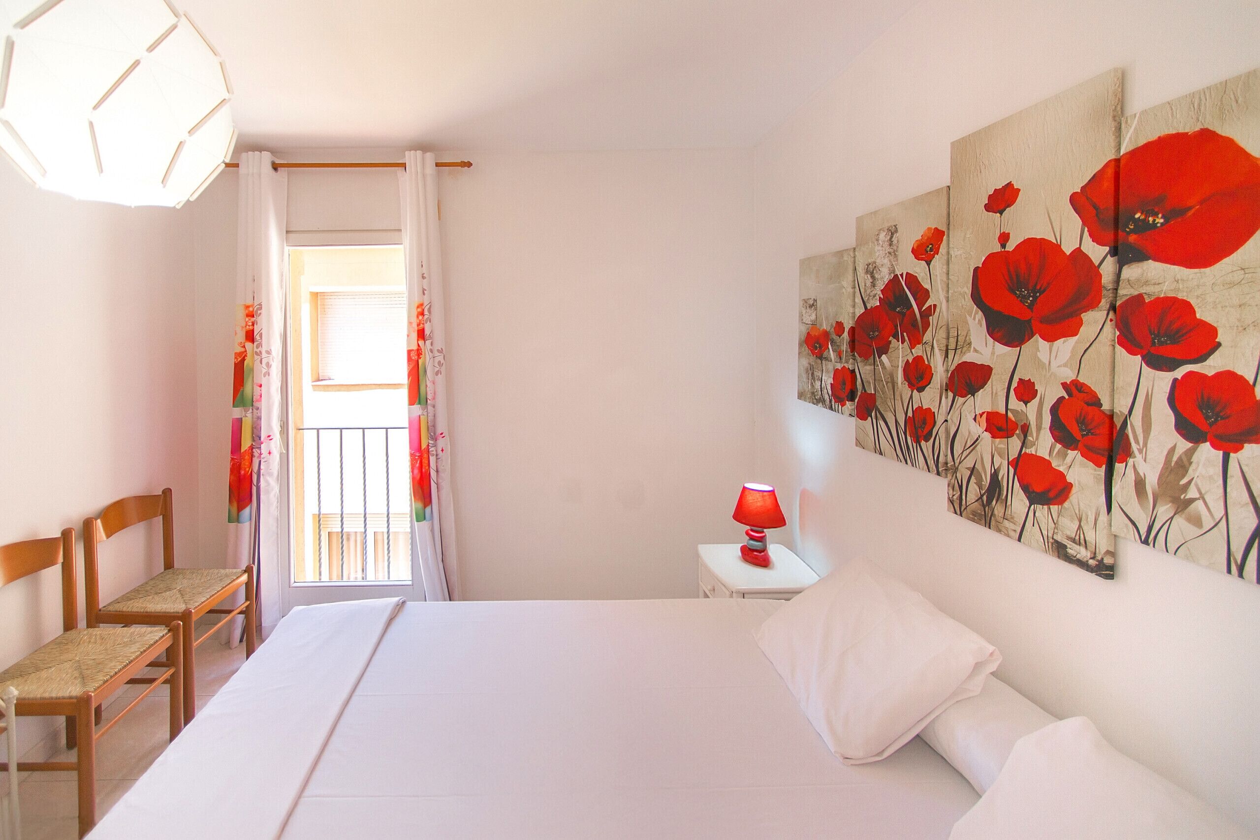 Una camera da letto con un letto, due sedie di legno, un comodino con una lampada rossa e un grande quadro floreale appeso alla parete.