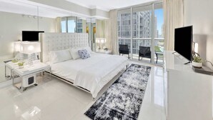 A modern hotel bedroom with a large bed, a TV, a desk, and a balcony with a city view.