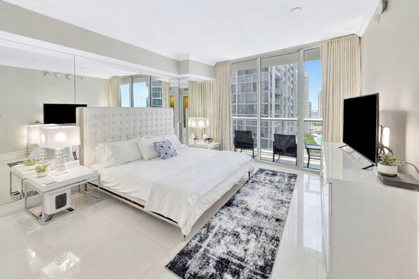 A modern hotel bedroom with a large bed, a TV, a desk, and a balcony with a city view.