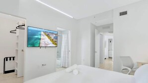 A modern hotel room with a large bed, a flat-screen TV mounted on the wall, and a view of a beach through the window. - Chic 2BR Edgewater Condo w/ Views + Balcony (Miami)