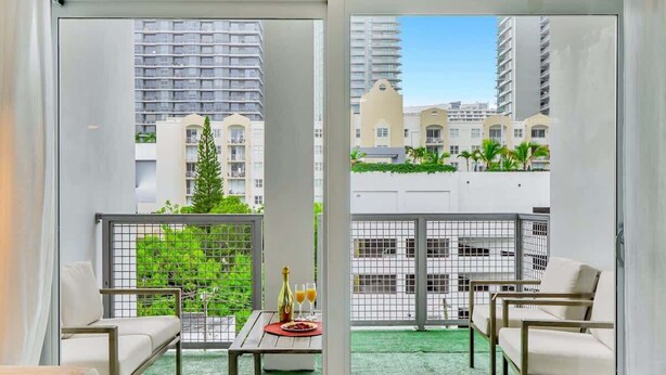 A balcony with a view of high-rise buildings, a table with drinks, and two chairs. - Chic 2BR Edgewater Condo w/ Views + Balcony (Miami)