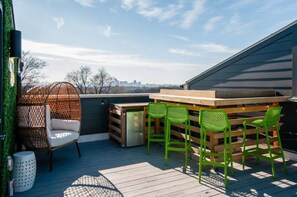 A deck with a wooden table and chairs, a wicker chair, and a trash can. - East Nash Lux - 13 Beds City Views (Nashville)