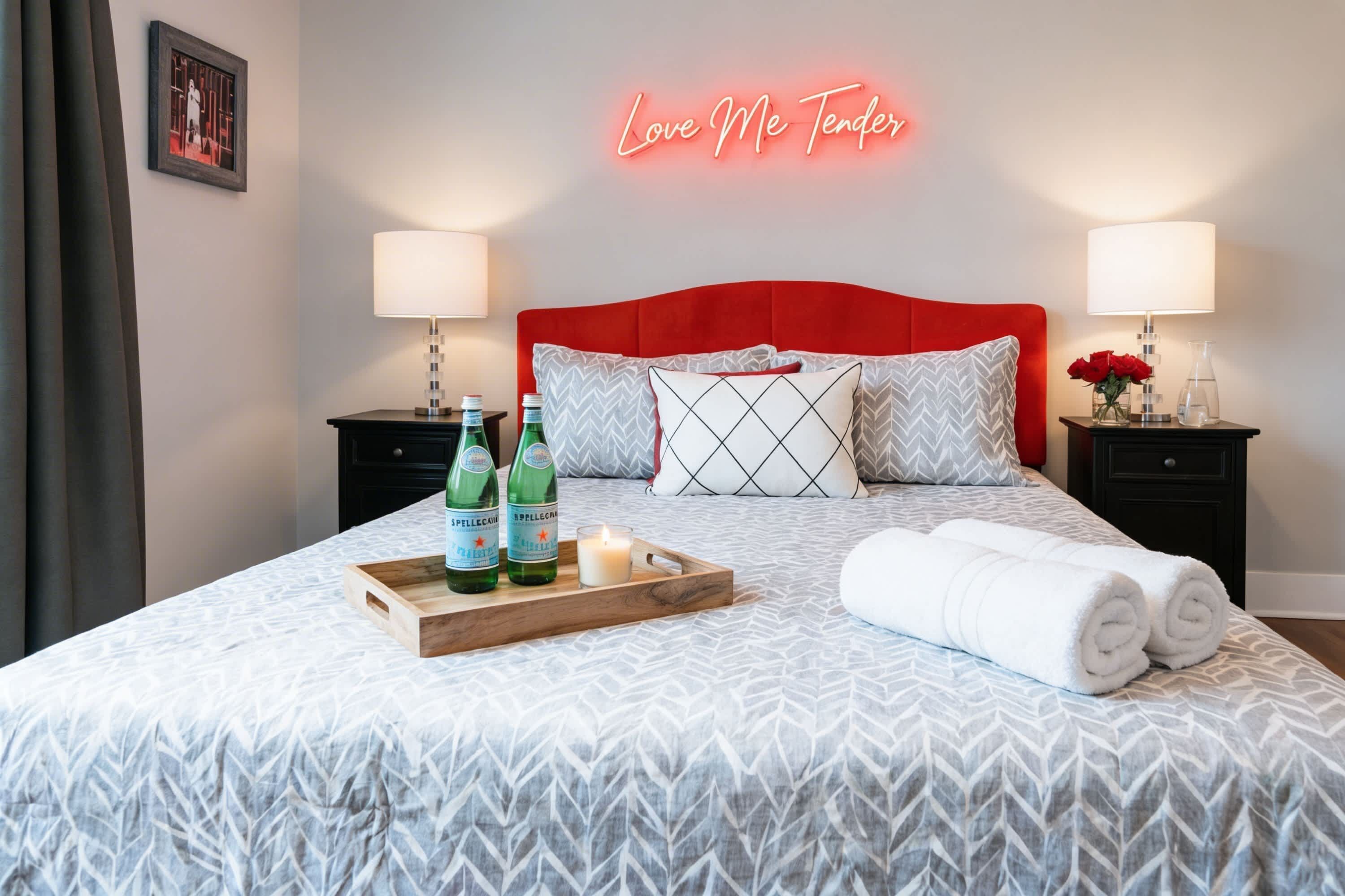 A neatly made bed with a red headboard, two bedside tables with lamps, a tray with bottles and a candle, and a neon sign above the bed.