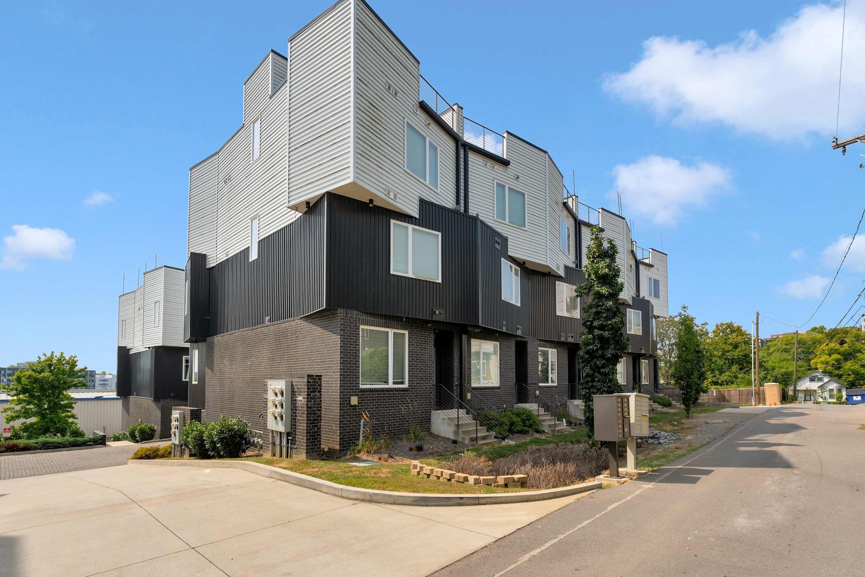 Un immeuble résidentiel moderne à plusieurs étages, avec un mélange de revêtements sombres et clairs, une entrée principale bien visible et un petit jardin à l’avant, agrémenté d’aménagements paysagers.
