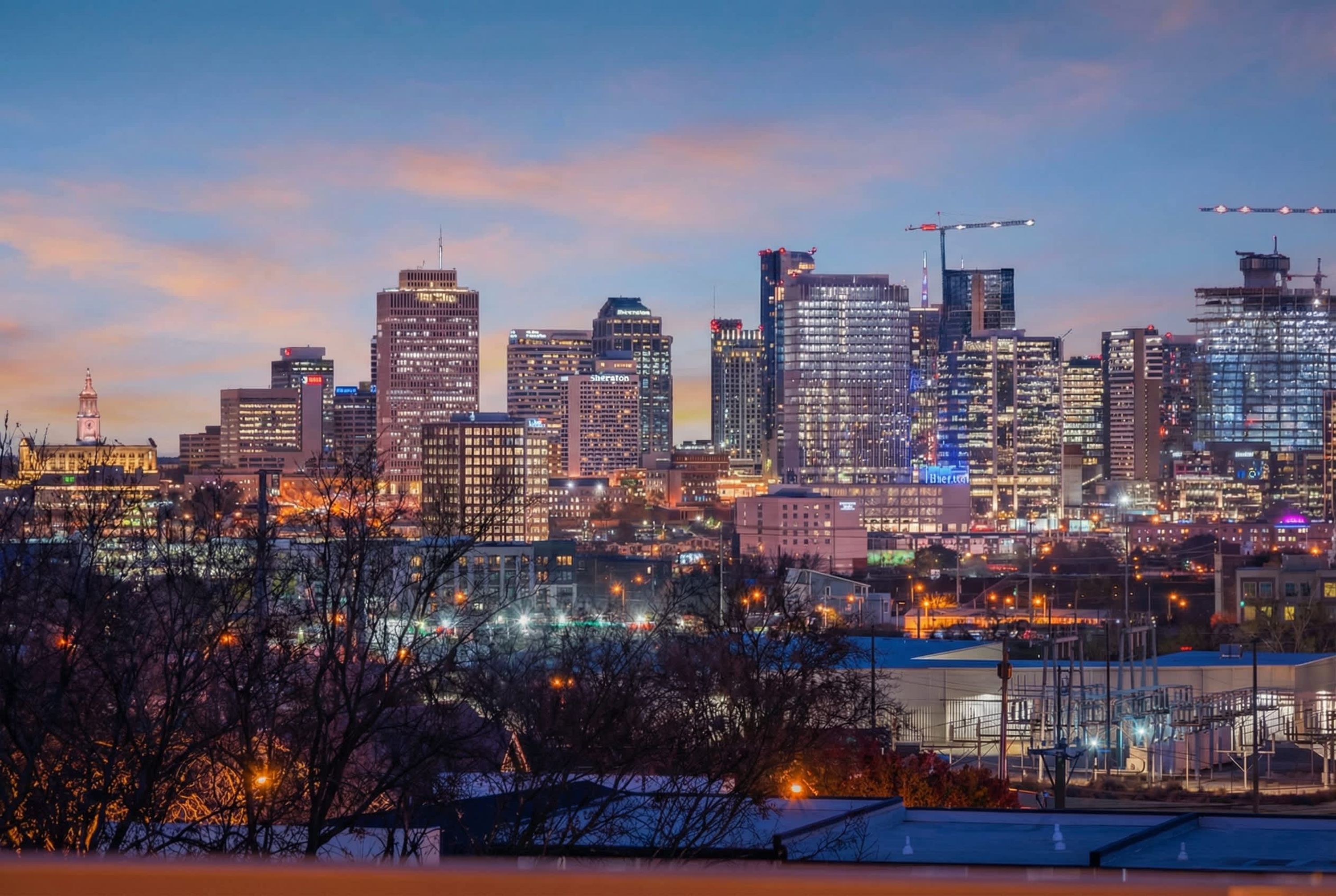 A city skyline at dusk with illuminated buildings and construction cranes.