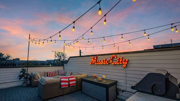 An outdoor patio with string lights, a neon sign reading "Music City," wicker furniture, and a grill.