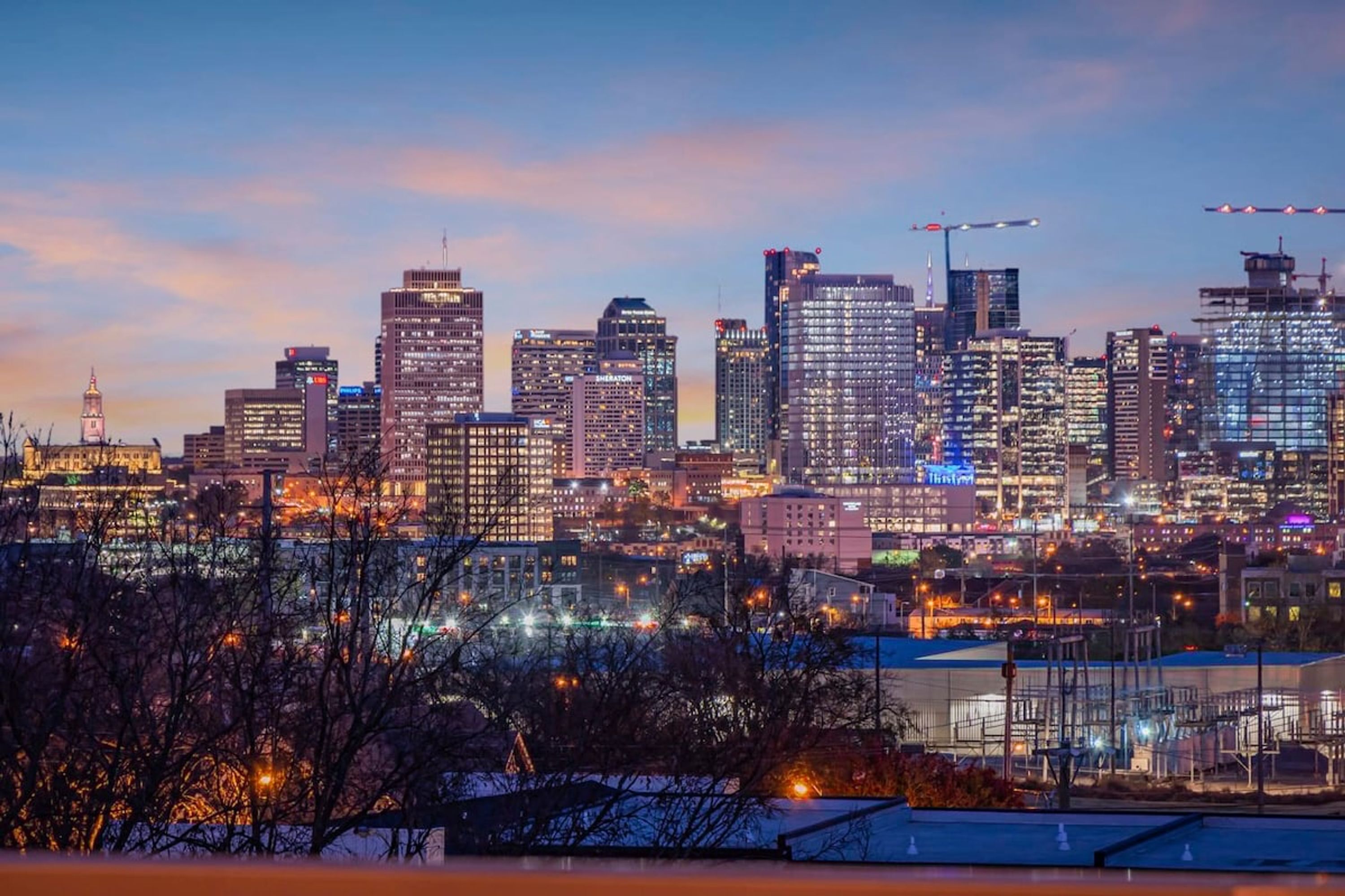 A city skyline at dusk with illuminated buildings and construction cranes.
