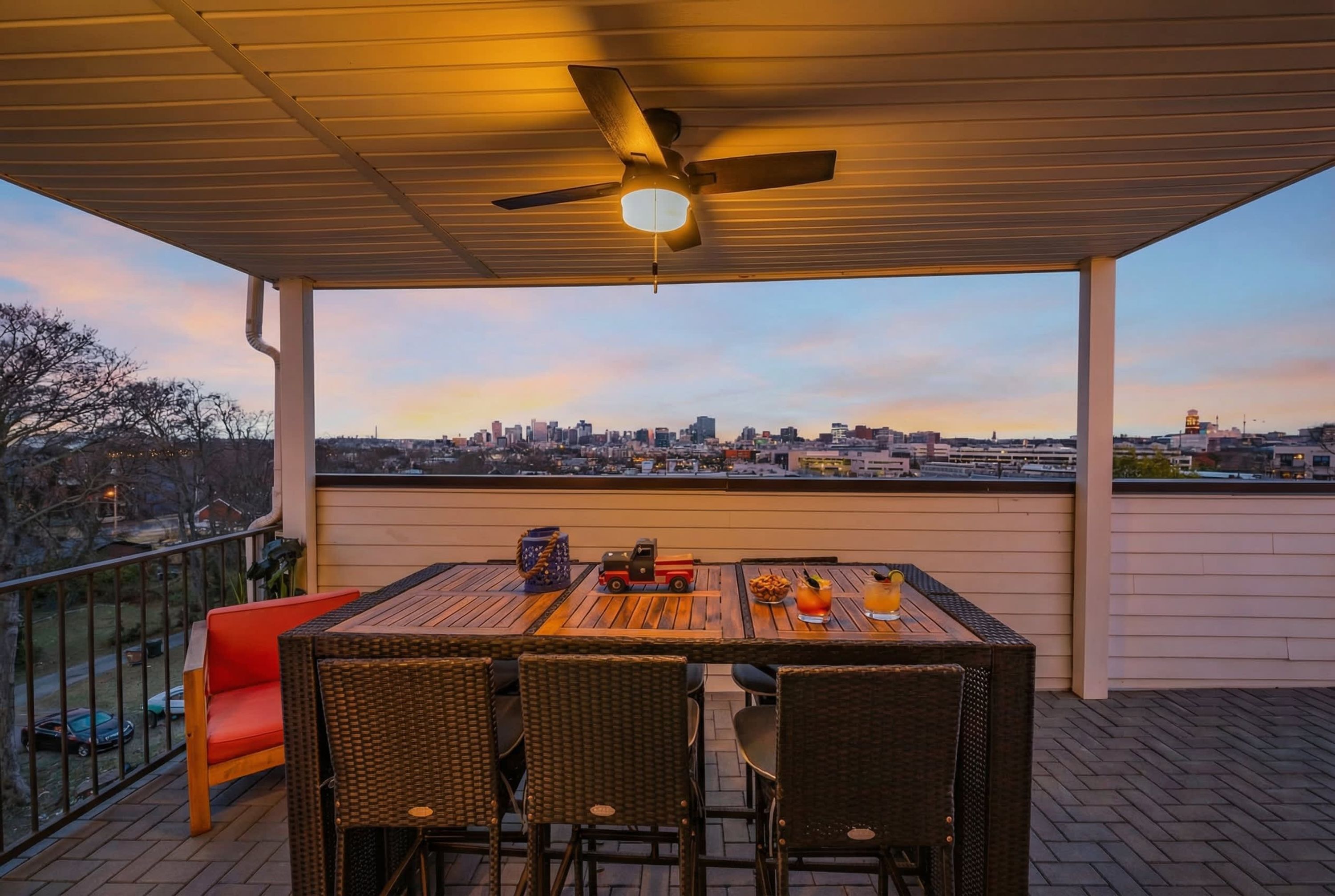 Eine überdachte Terrasse mit Tisch und Stühlen, einem Deckenventilator und Blick auf eine Stadtlandschaft.