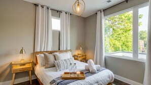 A bedroom with a bed, bedside tables, a nightstand, a window with curtains, and a hanging light fixture.