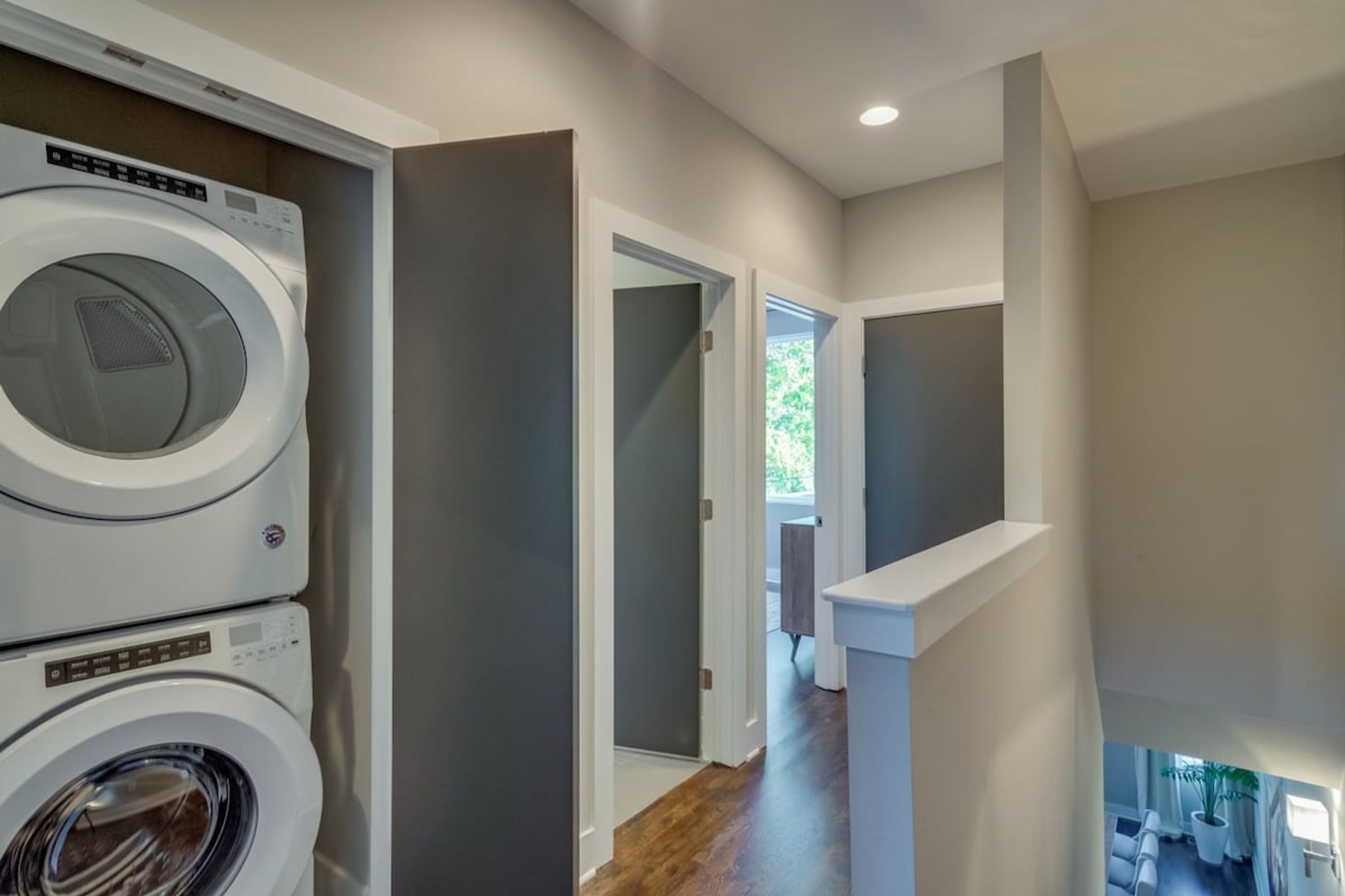 A laundry room with stacked washing machines, a hallway leading to a bedroom, and a view of a balcony with a potted plant.