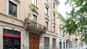 A building with a closed shop front, a balcony with plants, and a street with parked cars. - GuestHost - Island Cosy Pink Studio Apt (Milano)