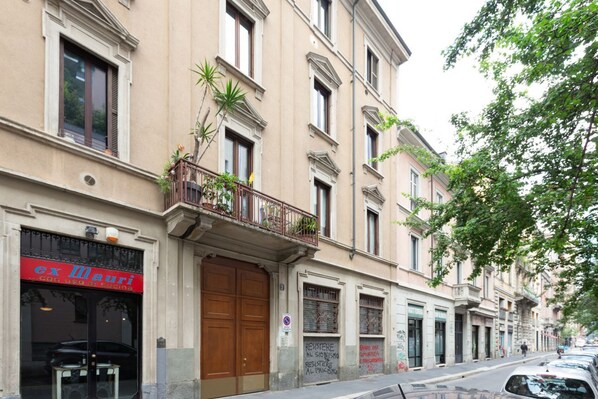 A building with a closed shop front, a balcony with plants, and a street with parked cars.