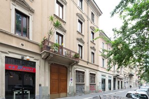 Un edificio con escaparate cerrado, un balcón con plantas y una calle con coches estacionados.