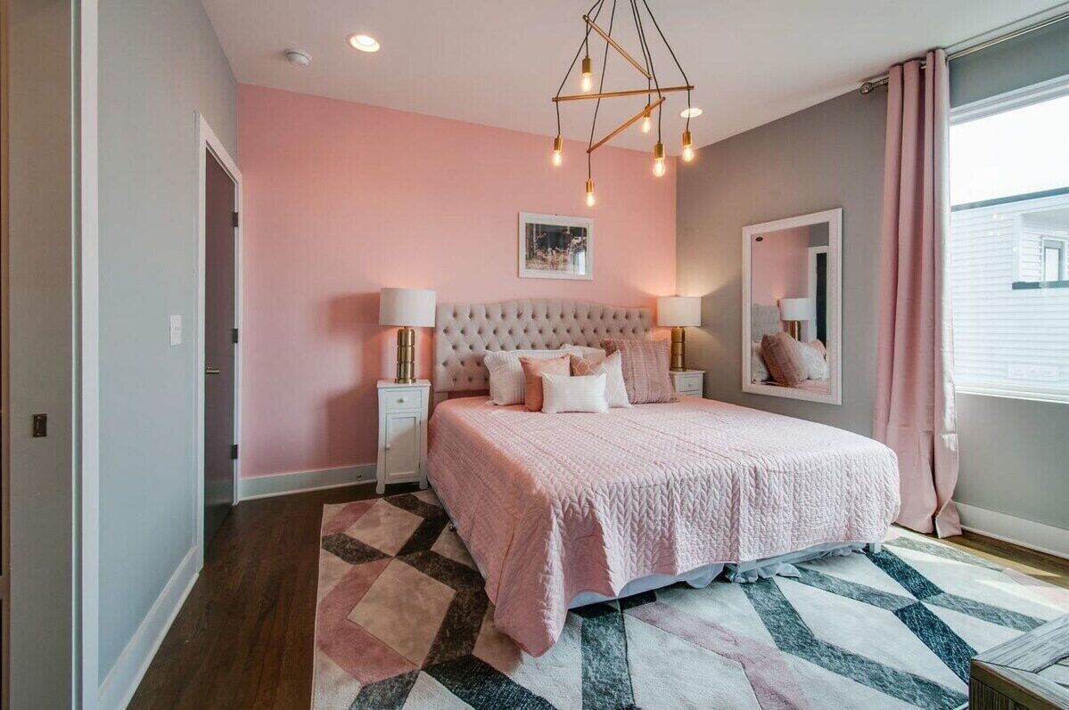 A bedroom with a tufted headboard, a pink bedspread, bedside lamps, and a geometric rug.