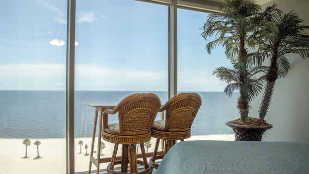 A room with a large window overlooking a beach, two wicker chairs, a potted palm tree, and a bed with a patterned bedspread.
