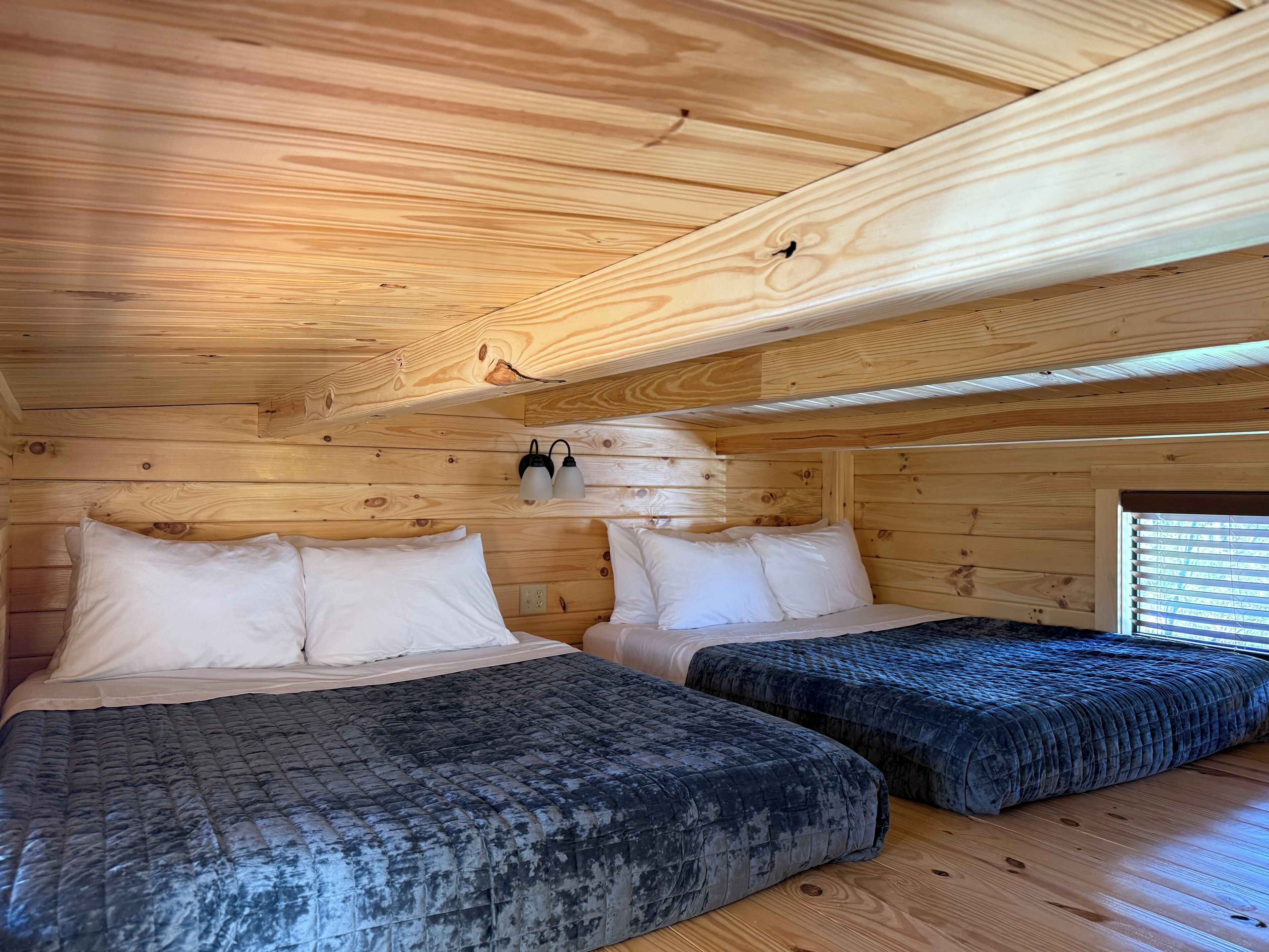 A cabin room with two beds, wooden walls, a sloped ceiling, and a window with blinds.