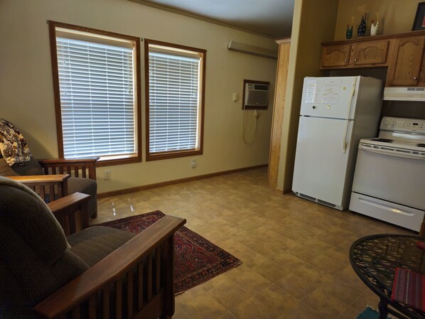 Una sala de estar con un sofá, una mesa de madera y un tapete estampado. Hay un área de cocina con refrigerador, estufa y armarios.