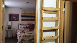 A room with a wooden ladder, a bed with patterned bedding, a small bedside table with a lamp, and a bathroom with a sink.