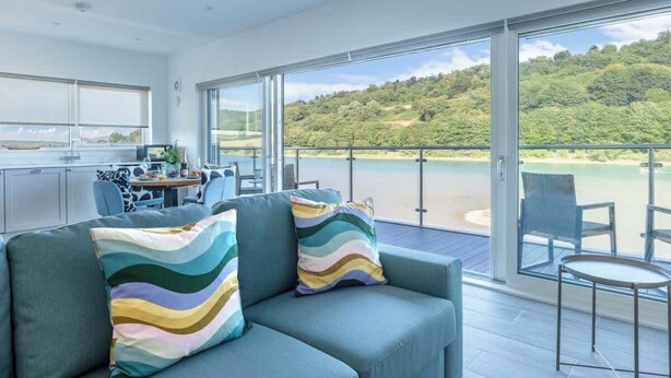Een moderne woonkamer met een bank en fauteuils, een eethoek met een tafel en stoelen, en een balkon met uitzicht op een strand en bos.