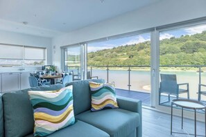 A modern living room with a sofa and chairs, a dining area with a table and chairs, and a balcony with a view of a beach and forest. - River View Prime River Front Location (Seaton)