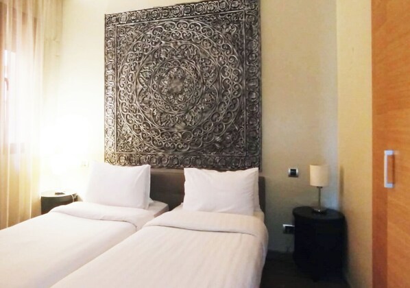 A hotel room with two beds, a large decorative tapestry on the wall, a nightstand with a lamp, and a wooden door.