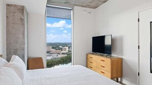 Une chambre avec une grande fenêtre, un lit, un téléviseur posé sur une commode en bois, et une vue sur des immeubles et de la verdure.