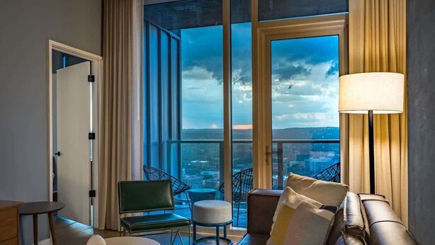Une chambre d’hôtel moderne dotée d’une grande fenêtre, d’un canapé, d’un fauteuil, d’une lampe et d’une petite table sur laquelle se trouve un plateau.