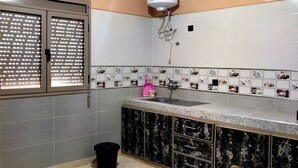 Une cuisine de taille réduite, équipée d’un évier, d’une fenêtre avec une grille métallique, d’un chauffe-eau fixé au plafond et d’un pare-feu en carrelage.