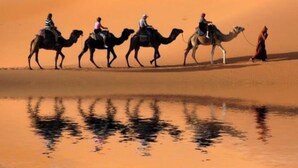 Un groupe de personnes chevauche des chameaux à travers un paysage désertique, avec des dunes de sable et une étendue d’eau qui reflète la scène.