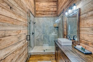 Ein Badezimmer mit Holzwänden, einer gläsernen Duschkabine, einem weißen Waschbecken und einem Spiegel mit Beleuchtung.
