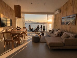 Un salon moderne doté d’une grande fenêtre donnant sur un plan d’eau, un coin repas avec des meubles en bois et un espace salon confortable.