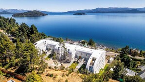 A modern building with multiple levels and a rooftop terrace, surrounded by dense greenery and overlooking a large body of water with mountains in the distance. - B612 Bariloche (San Carlos de Bariloche)