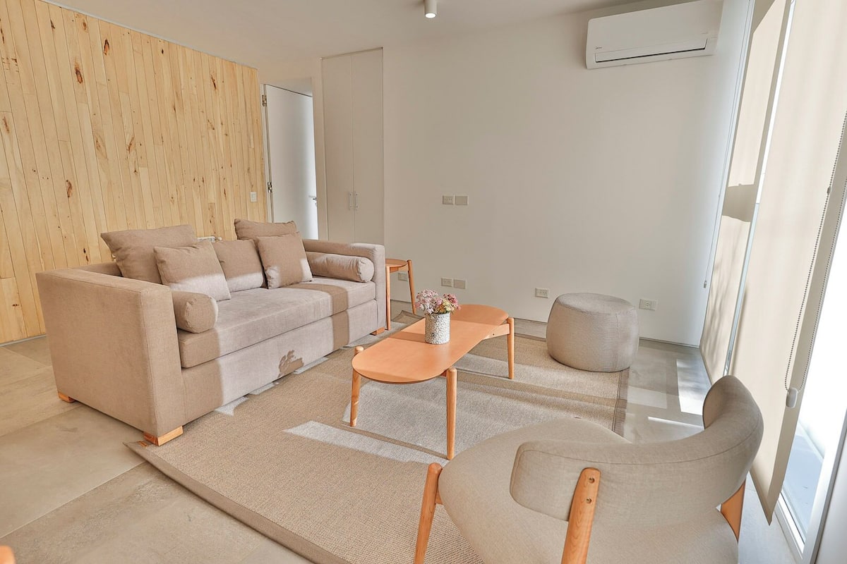 Uma sala de estar moderna com um sofá bege, uma mesa de centro de madeira e um banquinho redondo.