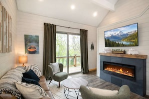 A cozy living room with a fireplace, a comfortable sofa, a TV mounted above the fireplace, and a large window with curtains.