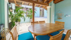Una mesa de comedor de madera con cojines azules, rodeada de sillas de madera, en una habitación con suelo de baldosas y techo de madera.
