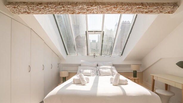 A bedroom with a skylight, a bed with white linens, a bedside table with a lamp, and a view of the city through the windows. - Bright & Chic Duplex with A/C – Heart of Paris 9th (Paris)