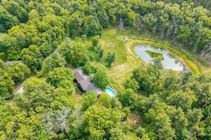 An aerial view of a property with a house, a swimming pool, and a pond surrounded by dense forest.