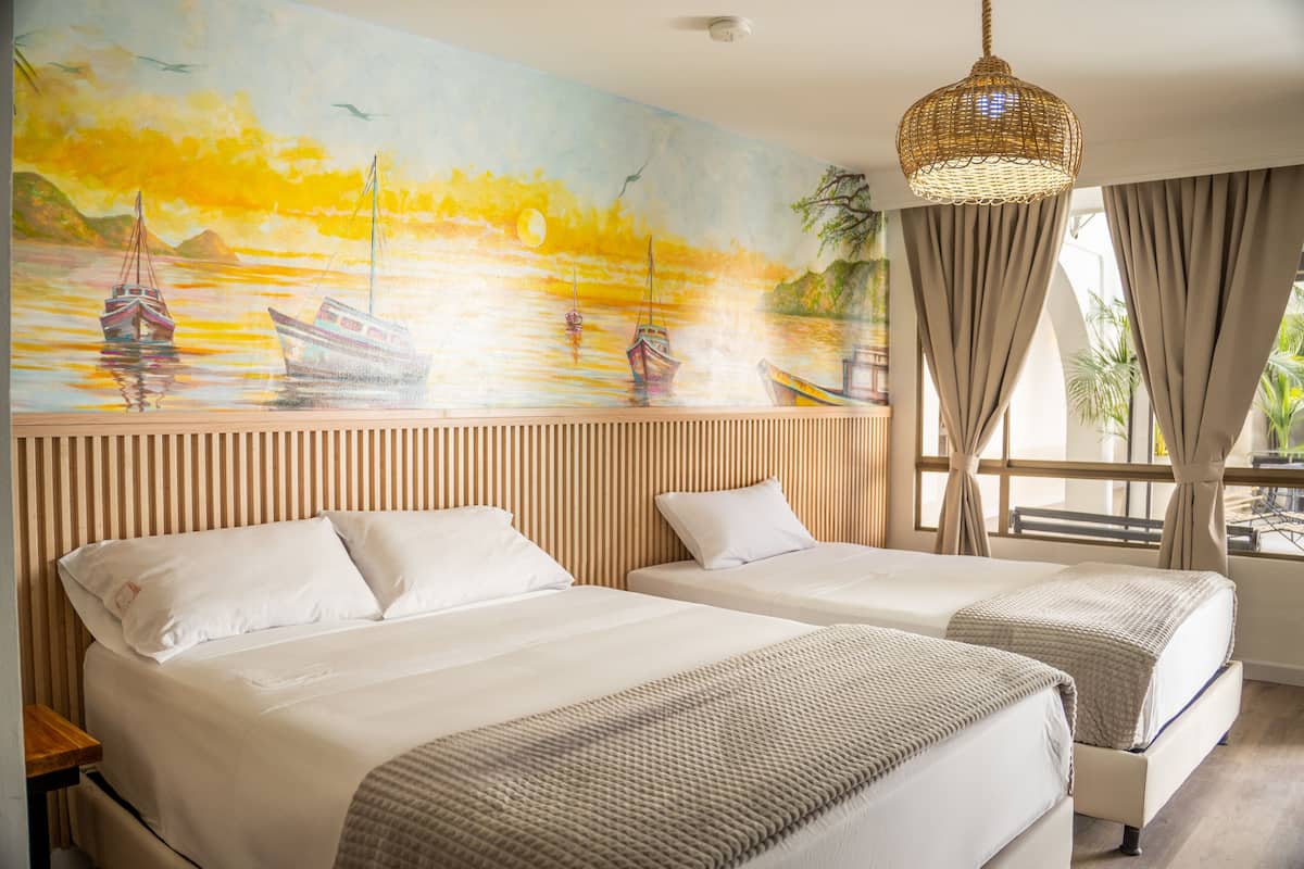 A hotel room with two beds, a large painting of boats on water, a woven light fixture, and a view of the outdoors through a window.