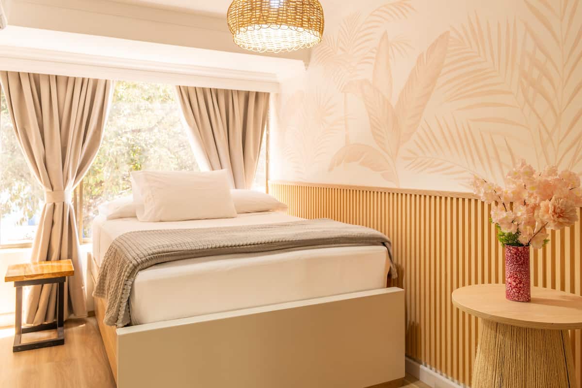 A bedroom with a bed, curtains, a small table, a round side table with a vase, and a wall with a palm leaf pattern.