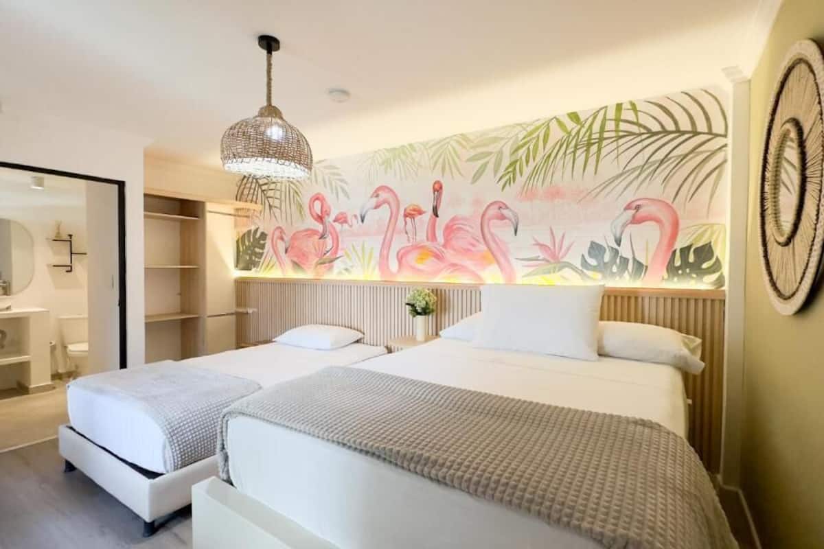 A hotel room with two beds, a large wall mural of flamingos, a hanging light fixture, and a wooden headboard.
